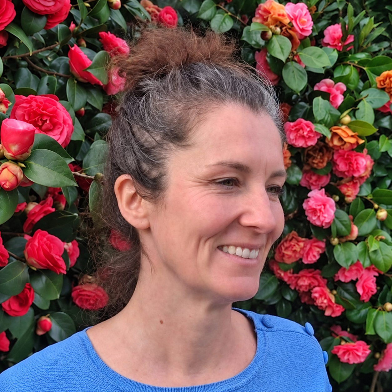 Marine Croiset souriante, en pull bleu, devant des fleurs rouges et du feuillage.