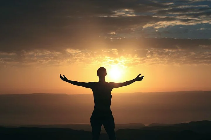 Silhouette d’une personne les bras ouverts face au soleil, devant un paysage de collines.