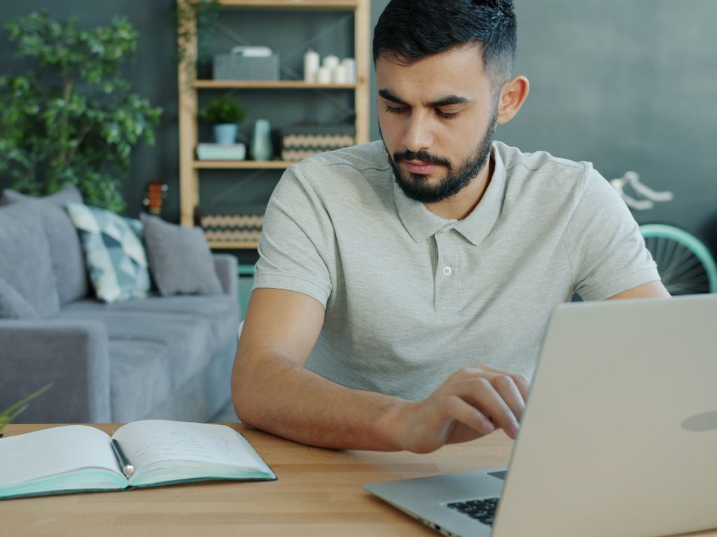 Homme assis à une table devant un ordinateur portable, avec un carnet ouvert à ses côtés.